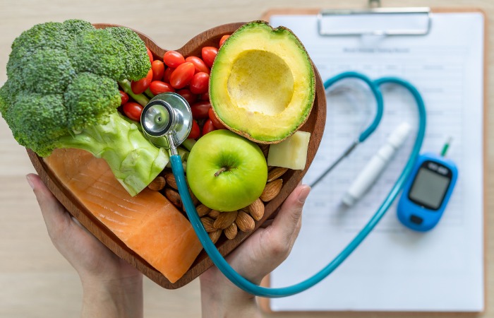 A heart-shaped bowl filled with broccoli, salmon, tomatoes, nuts, and apple, with stethoscope coming out of the bowl