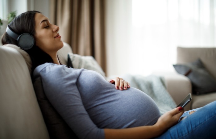 Pregnant woman sitting on couch while holding her phone and listening to music with headphones