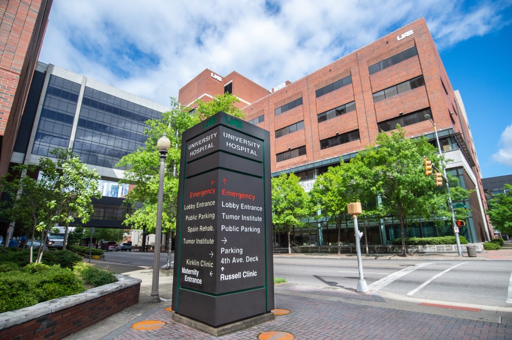 UAB University Hospital signage on sidewalk with the O'Neal Comprehensive Cancer Center, the North Pavilion, and blue sky and white clouds in the background, April 2020.