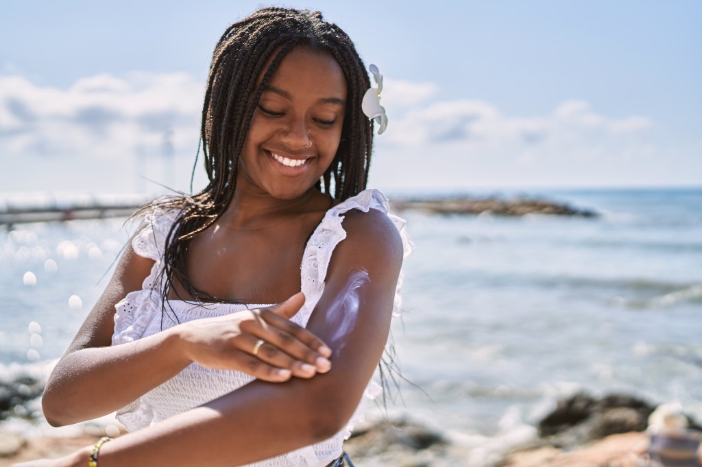 Young African American girl smiling while applying sunscreen at the beach.