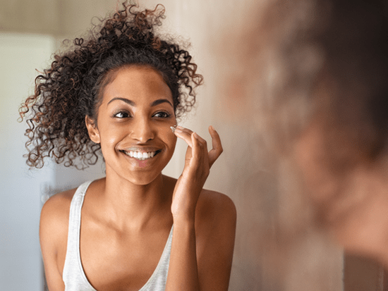 African American female smiling at herself in the mirror while applying moisturizer to her face