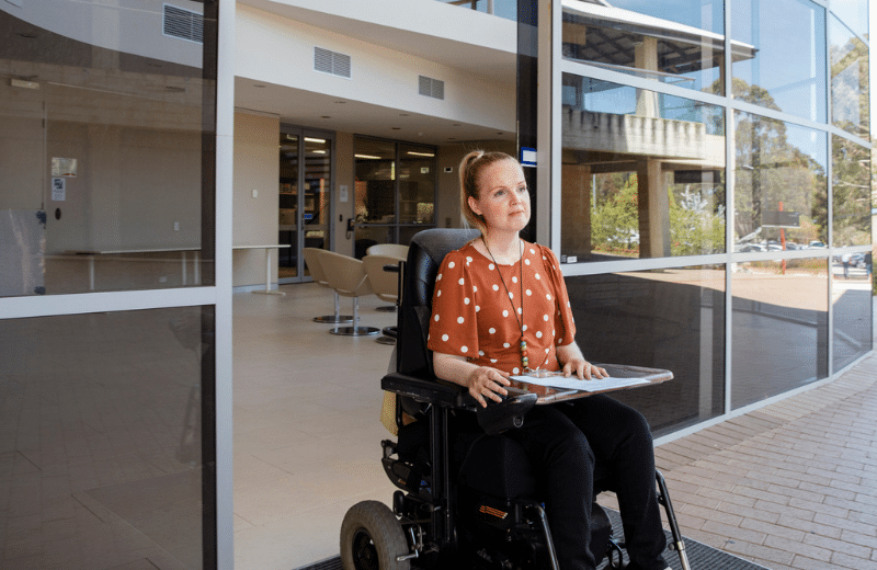 A front view of a woman leaving her office in her electric wheelchair take some paperwork to a colleague.
