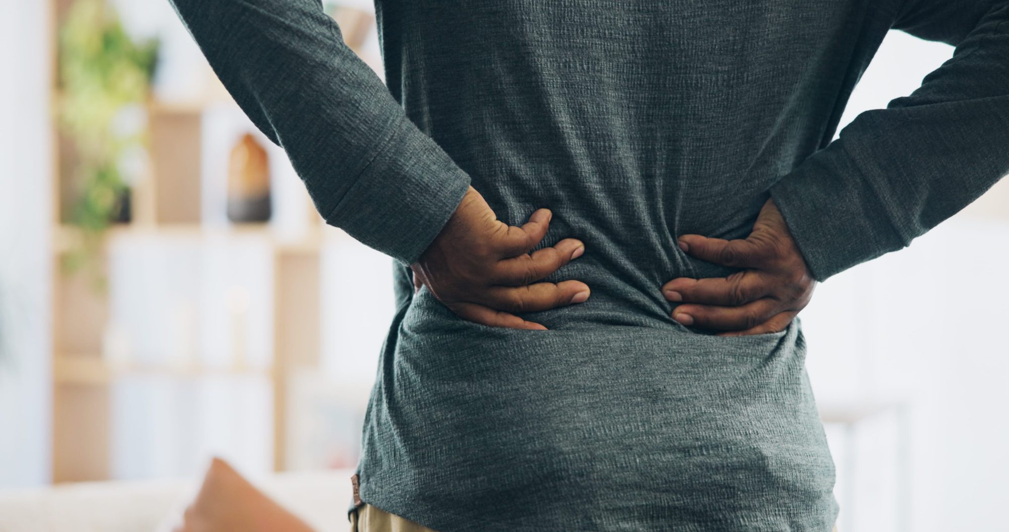 Black male holding lower back with both hands, indicating he has muscle tension or back pain while at home.