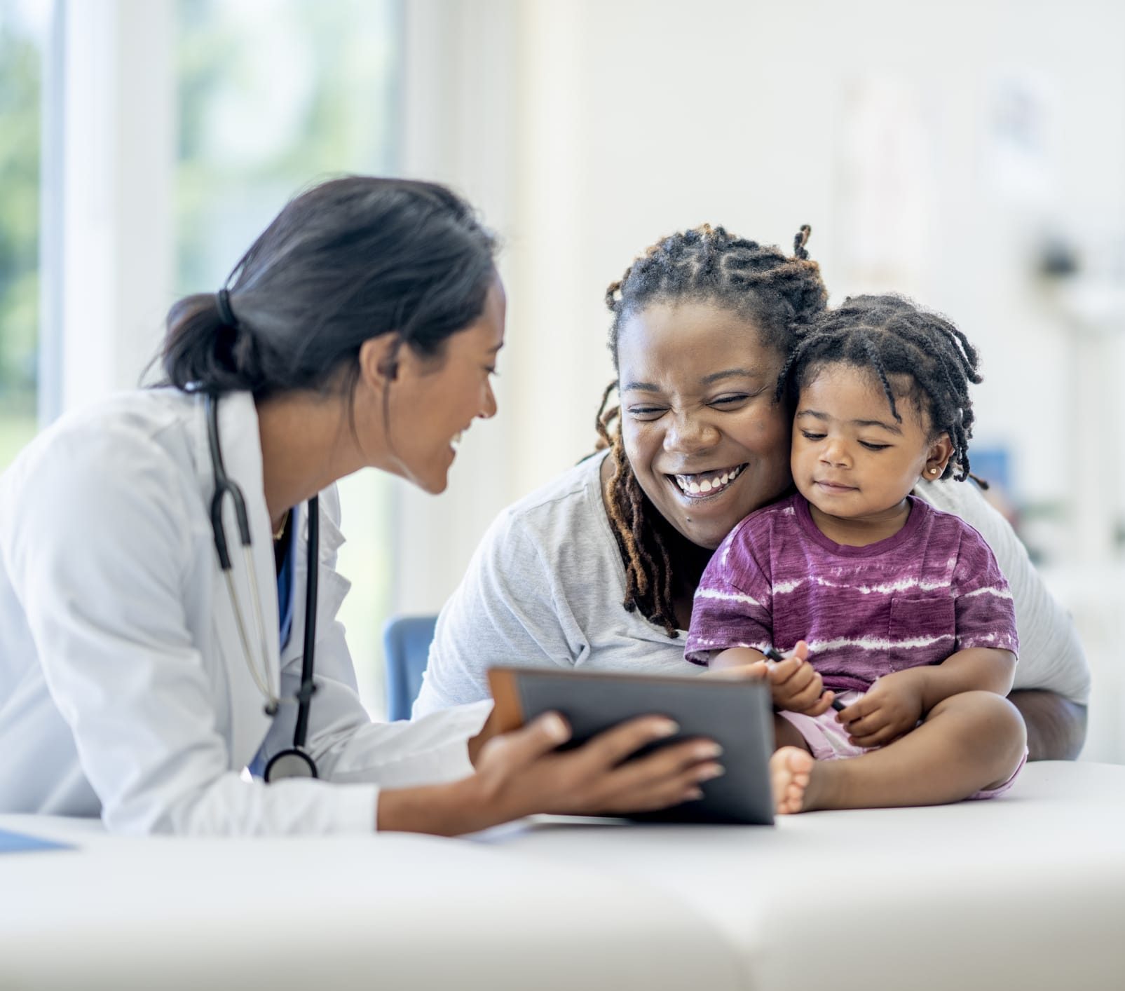 A young mother and her son visit the doctor together. The baby boy is seated on the exam table with his mother seated in front of him and making sure he does not fall.