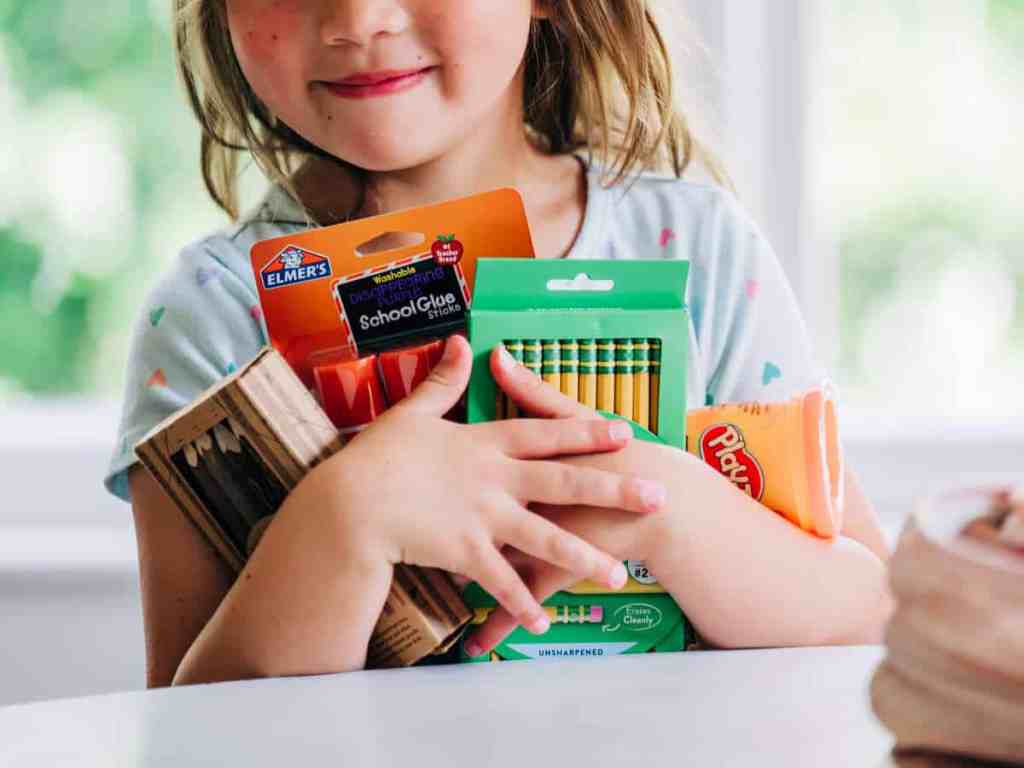 Close-up of smiling girl holding new school supplies, including pencils, crayons, and glue sticks, while standing beside dining table in home