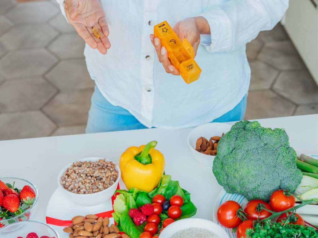 older woman holding a medicine organizer with vegetables on a table below